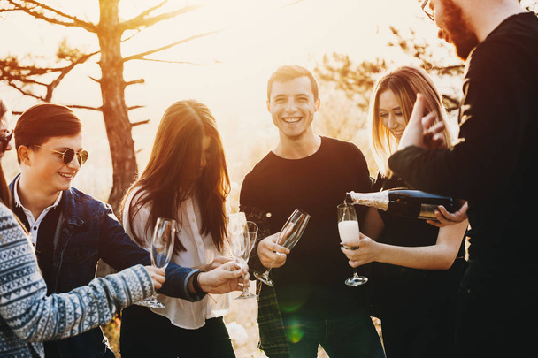 Группа привлекательных молодых людей смеется и разливает прекрасное шампанское, стоя на красивой природе во время вечеринки
 - Фото, изображение