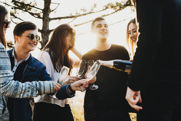 Unrecognizable guy pouring fine champagne into glasses of cheerful friends while celebrating holiday in countryside - Photo, Image