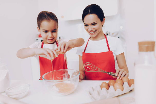 Mother Preparation Egg for Sweet Fod. Smile with Beautiful Daughter. Girl Happy with Mother when Mother Cook Sweet Cake. Red Apron for Mother and Daughter. Happy Mother with Happt Young Daughter. - Foto, Bild