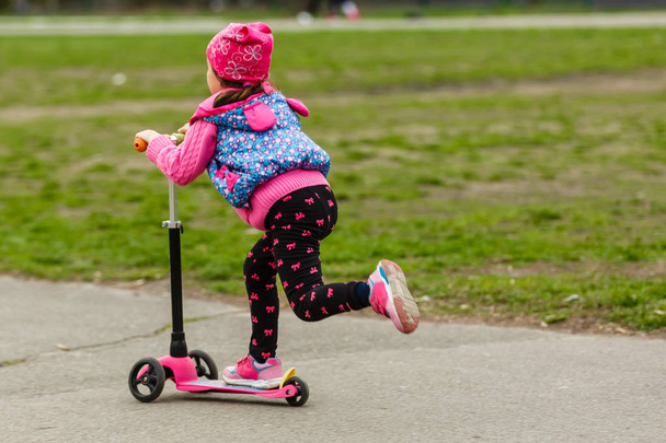 маленька дівчинка в рожевому пальто верхи скутер в парку
 - Фото, зображення