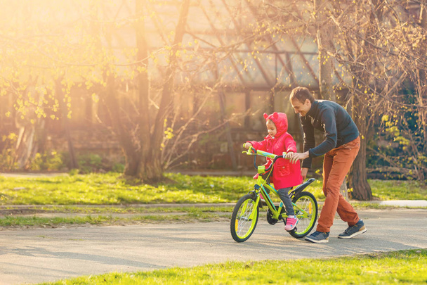 緑の公園で娘に続く自転車と幸せな父親に乗って小さな女の子 - 写真・画像