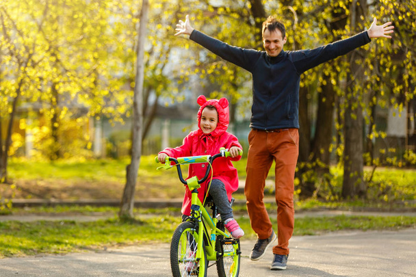 Menina andando de bicicleta e feliz pai seguindo filha no parque verde
 - Foto, Imagem