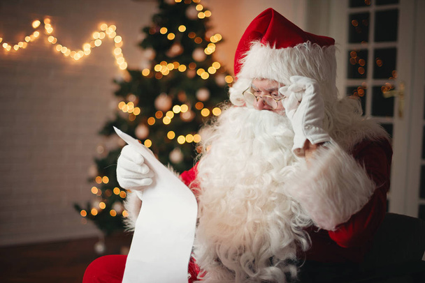 Santa Claus reading from a long list, in a cozy room   - Valokuva, kuva