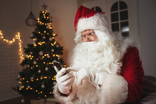 Santa Claus using a phone, sitting in a cozy room  - Фото, изображение