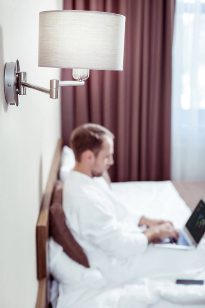 Blonde-haired man wearing white bathrobe lying on bed with laptop - Photo, Image