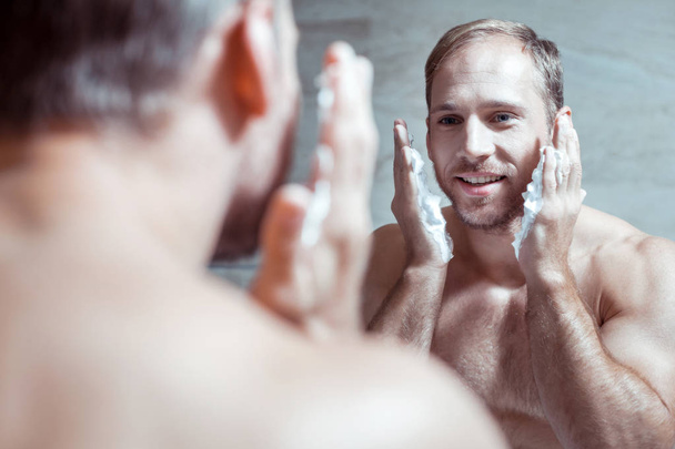 Cheerful blue-eyed man smiling while looking into mirror and shaving face - Фото, изображение