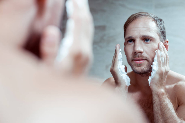 Handsome blue-eyed man using shaving foam while shaving face - Foto, Imagem