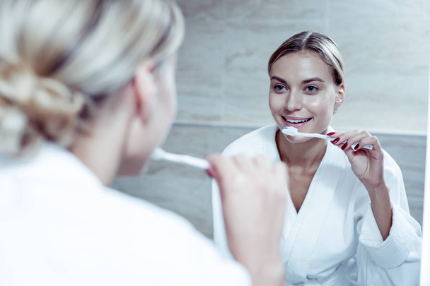 Beautiful blonde-haired woman wearing bathrobe brushing her teeth - Zdjęcie, obraz