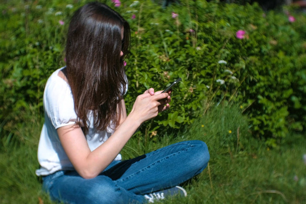 Woman is typing a message on mobile phone sitting in park in sunny day, chatting. Side view. - Photo, image