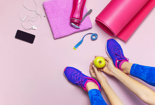 Fiatal nő kék cipők tart alma a kezében, előkészítése képzés. Üveg víz, jóga szőnyeg, telefon, fejhallgató, pasztell rózsaszín háttér lapos feküdt felülnézet. Sport cipő Fitness koncepció - Fotó, kép