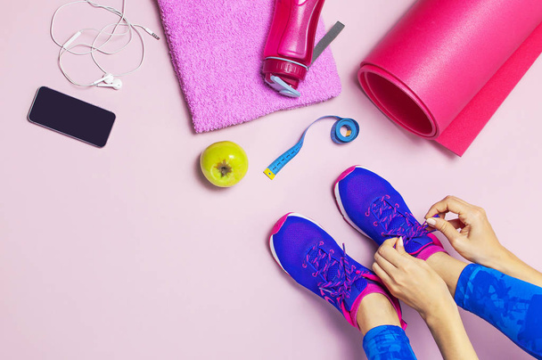 Young woman in blue sneakers holds apple in his hands, preparing for training. Bottle of water, yoga mat, phone, headphones on pastel pink background flat lay top view. Sports shoes Fitness concept - Foto, imagen