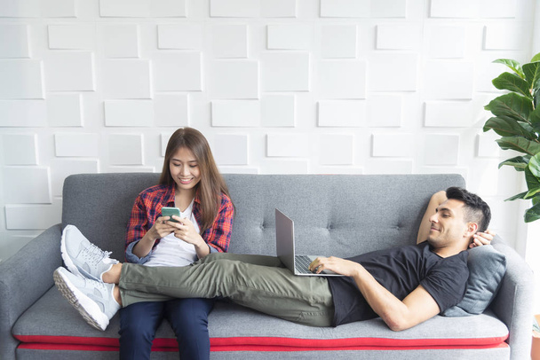 Ζευγάρι χαλαρώνοντας στον καναπέ στο σπίτι. Χρησιμοποιώντας το κινητό και το laptop για να παίξετε, να εργαστούν και να συνδεθείτε σε άλλο τους ανθρώπους στα social media. Σύγχρονη τεχνολογία για την ζωή. - Φωτογραφία, εικόνα