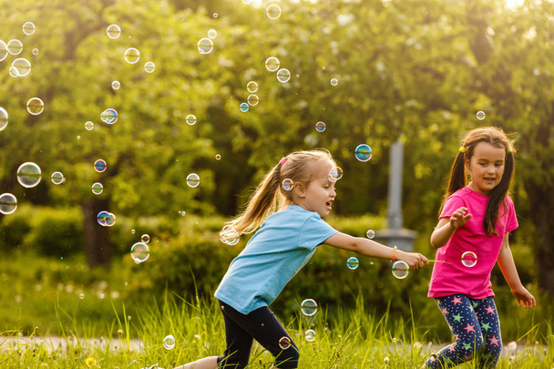 Duas meninas felizes sobre bolhas de sabão flutuantes no parque verde ao pôr do sol
 - Foto, Imagem
