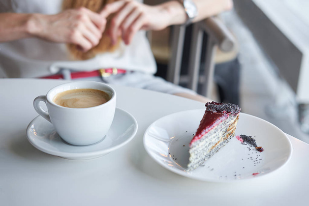 Чашка кофе и кусок пирога с маком на белом столе в кафе. Женщина на заднем плане готова к завтраку
. - Фото, изображение