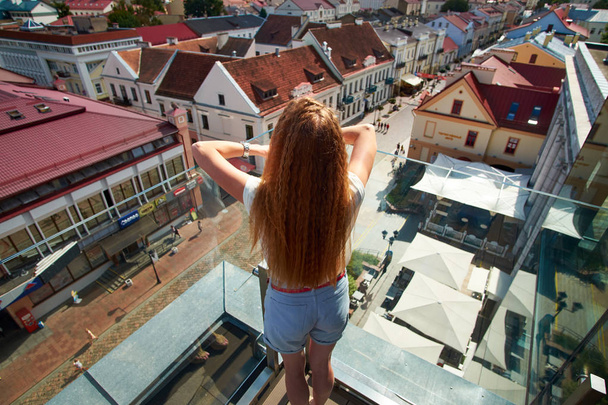 Σγουρό κορίτσι κοκκινομάλλα παρατηρεί ευρωπαϊκή πόλη από την οροφή - Φωτογραφία, εικόνα