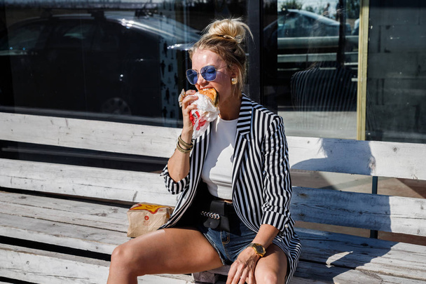 Portrait of lovely blonde girl wearing sunglasses, striped jacket and white t-shirt eating sandwich outdoors - Foto, Imagem