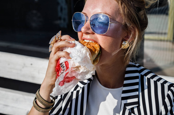Portrait of lovely blonde girl wearing sunglasses, striped jacket and white t-shirt eating sandwich outdoors - Photo, image