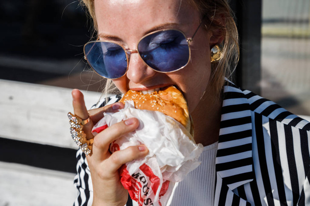 Portrait of lovely blonde girl wearing sunglasses, striped jacket and white t-shirt eating sandwich outdoors - Fotoğraf, Görsel