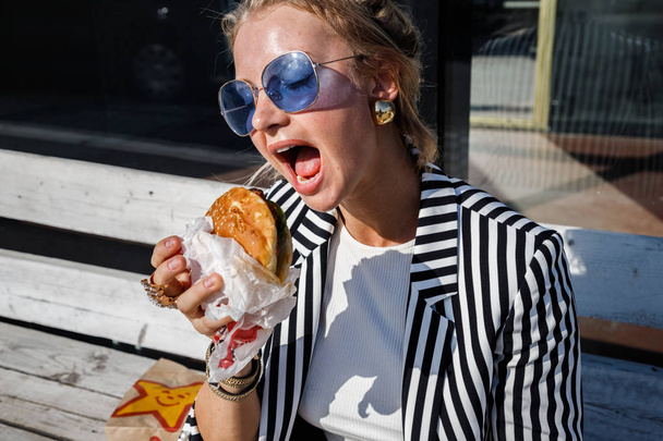 Portrait of lovely blonde girl wearing sunglasses, striped jacket and white t-shirt eating sandwich outdoors - Foto, Imagem