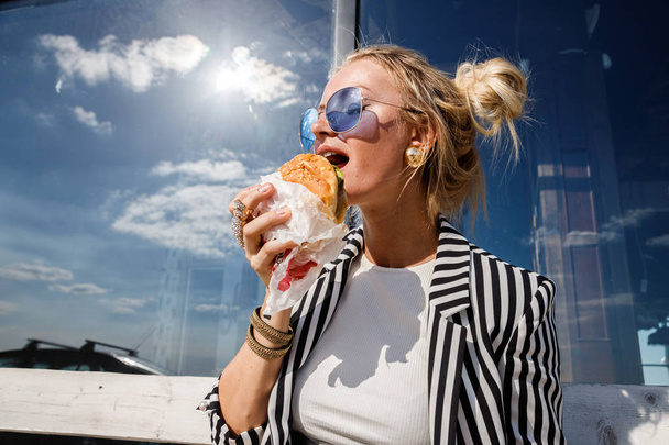 Portrait of lovely blonde girl wearing sunglasses, striped jacket and white t-shirt eating sandwich outdoors - Фото, зображення