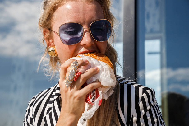 Portrait of lovely blonde girl wearing sunglasses, striped jacket and white t-shirt eating sandwich outdoors - Foto, afbeelding
