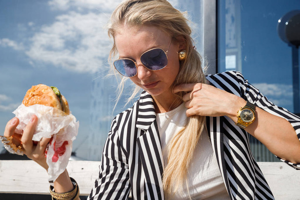 Portrait of lovely blonde girl wearing sunglasses, striped jacket and white t-shirt eating sandwich outdoors - Fotografie, Obrázek