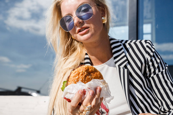 Portrait of lovely blonde girl wearing sunglasses, striped jacket and white t-shirt eating sandwich outdoors - Foto, Imagen