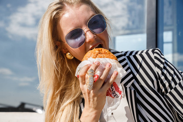 Portrait of lovely blonde girl wearing sunglasses, striped jacket and white t-shirt eating sandwich outdoors - Photo, Image