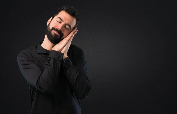 Handsome man with beard making sleep gesture on black background - Photo, Image