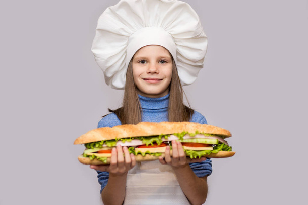 Ευτυχισμένο κοριτσάκι με hot dog και ομοιόμορφο σεφ που απομονώνονται σε λευκό φόντο. Παιδί σεφ. - Φωτογραφία, εικόνα