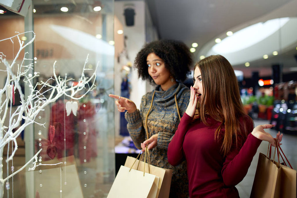 Young multiethnic diverse race women with paper shopping bags near decorated shopwindow. Girl pointing by finger at shopfront, clothes and products in mall on christmas new year sale. - 写真・画像