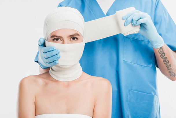 Nahaufnahme eines plastischen Chirurgen mit Handschuhen, der das Gesicht der Frau mit einem Verband isoliert auf weißem Grund abklebt - Foto, Bild