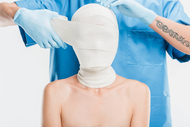 zblízka plastický chirurg v rukavicích nahrávat žena tvář s obvazem izolované na bílém - Fotografie, Obrázek