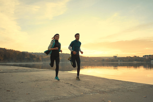 Par motivado de corredores para correr no lago ao nascer do sol. Jovem e mulher em roupas esportivas, correndo juntos. Pessoas esportivas treinando ao ar livre
 - Foto, Imagem