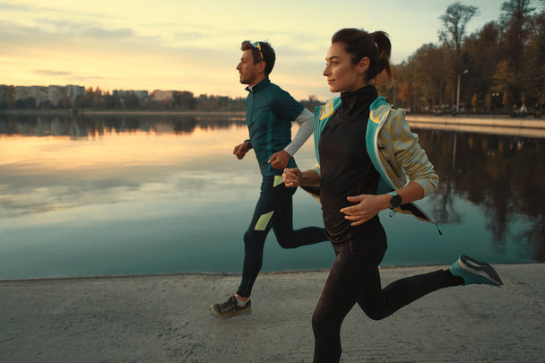 Un par de corredores motivados a correr por el lago al amanecer. Joven hombre y mujer en ropa deportiva, corriendo juntos. Formación de deportistas al aire libre
 - Foto, imagen