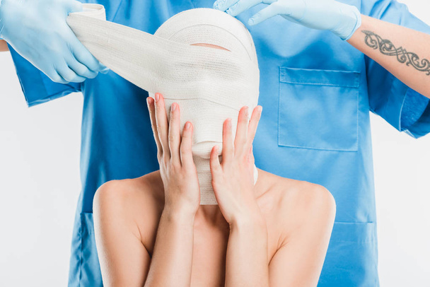 крупным пластмассовым хирургом в перчатках, заклеивающим лицо женщины повязкой, изолированной на белом
 - Фото, изображение
