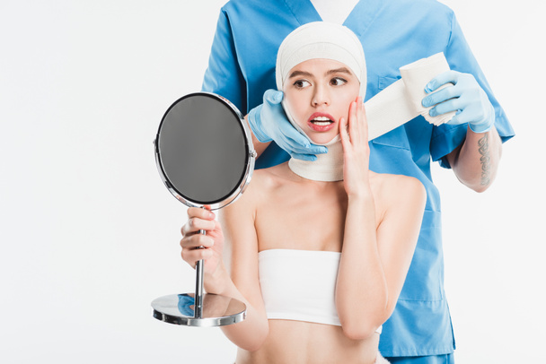 Chirurg in Handschuhen verklebt Gesicht mit Verband nach Schönheitsoperation, während verängstigte Frau auf weißen Spiegel schaut - Foto, Bild
