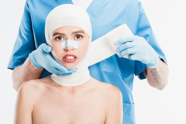  surgeon in gloves taping up woman face with bandage after plastic surgery isolated on white - Photo, Image
