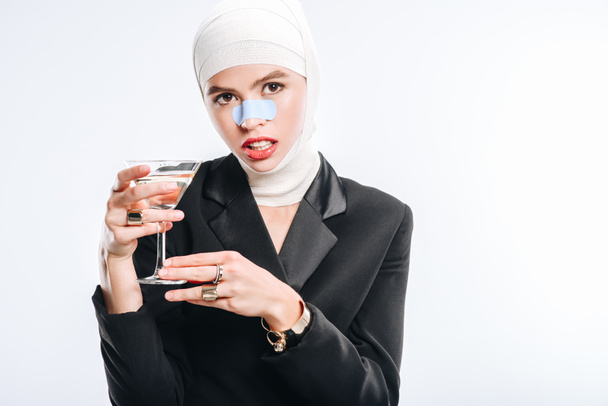 стильная женщина с бинтами на голове после пластической операции держа стекло с коктейлем изолированы на белом
 - Фото, изображение