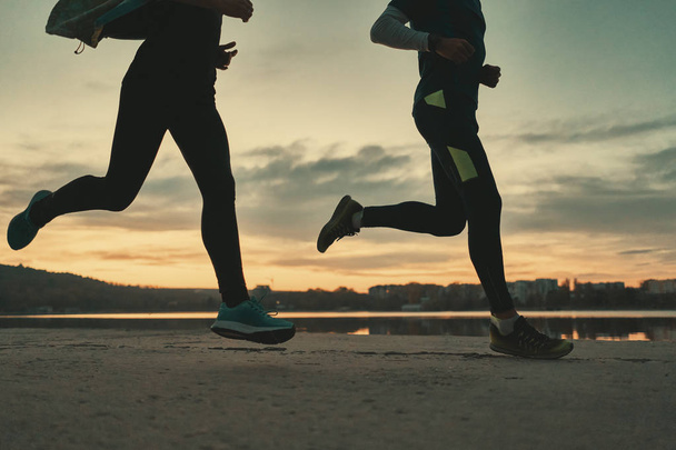 A fiatal pár futók lábakkal napfelkeltekor a tó és az ég, a háttérben futó. Sportos emberek edzéseket a szabadban végzi. Le szög lövés. - Fotó, kép