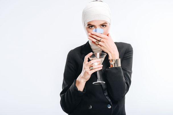 стильная женщина с бинтами на голове после пластической операции держа стекло с коктейлем и закрывая рот рукой изолированы на белом
 - Фото, изображение