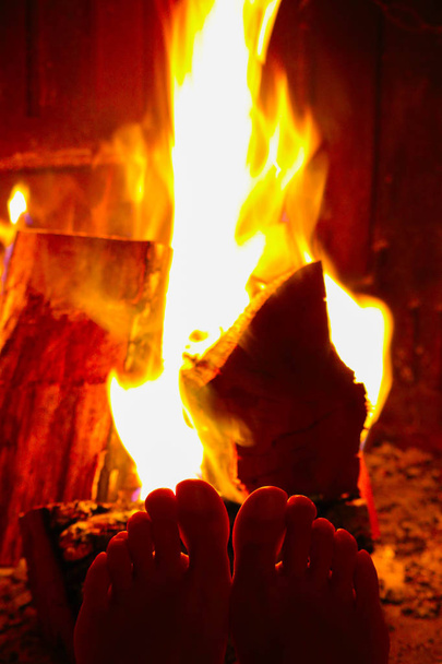 Burning wood at the fireplace, female legs is warming up. Firewood bricks at the fire, woman foot heating. - Photo, Image