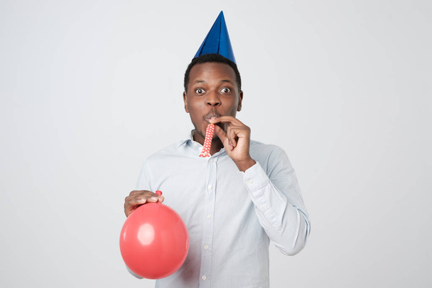 Jonge Afrikaanse man plezier op partij draagt blauwe shirt en vakantie hoed, partij hoorn blazen. - Foto, afbeelding