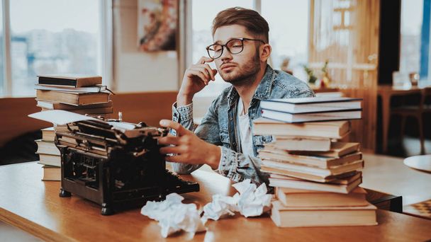Concept of Young Guy Working on Typewriter. Handsome Hardworking Freelance Screenwriter Scenarist Sitting at Table Near Stacks of Books and Crumpled Paper Sheets Writing Article, Novel, Play. - Valokuva, kuva