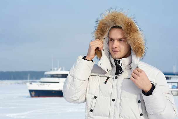 Fashion portrait of the young beautiful man in a white jacket with fur hood in winter - Foto, afbeelding