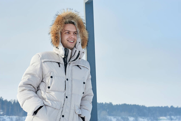 young beautiful man in a white jacket with fur hood in winter Siberia. Fashion portrait . - Фото, зображення
