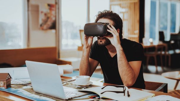 Happy Designer Working Remotely and Testing VR. Portrait of Cheerful Caucasian Guy Wears Virtual Reality Goggles Sitting at Desk Using Laptop ang Gadgets Holds Hand Up Indoors. - Photo, image