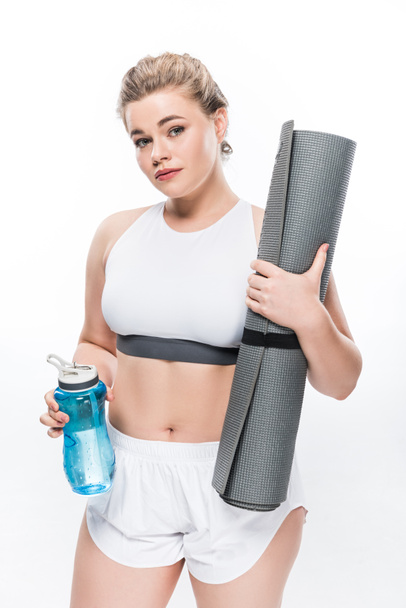 schöne übergewichtige Mädchen hält Flasche Wasser und Yogamatte isoliert auf weiß  - Foto, Bild