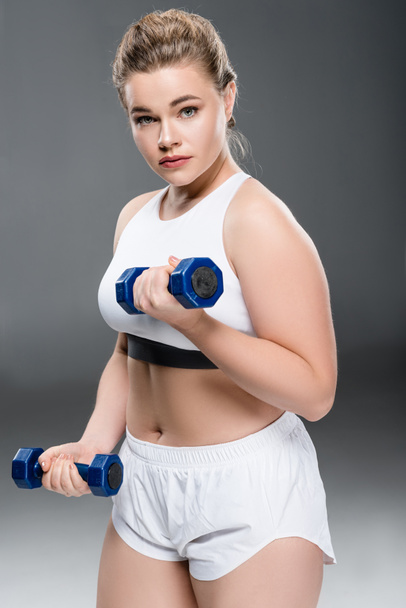 young size plus woman exercising with dumbbells and looking at camera on grey - Фото, зображення