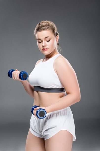 young size plus woman exercising with dumbbells and looking down on grey - Foto, Imagem